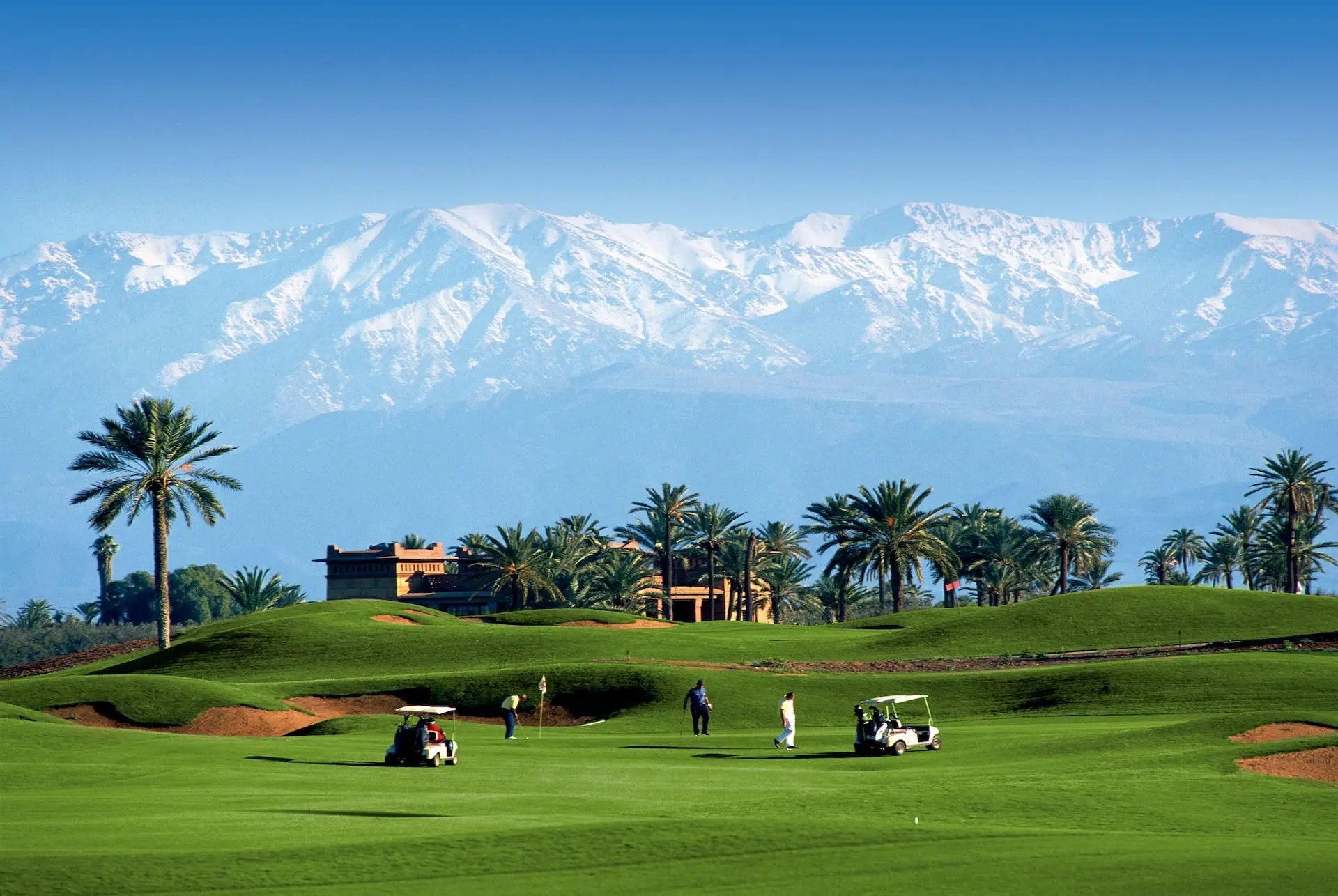 Golfers and buggies on a palm-lined Marrakech fairway with the snow-capped Atlas Mountains behind